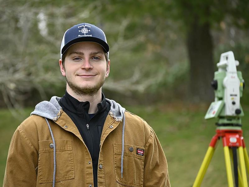A man wearing a baseball cap standing outside next to a piece of surveying equipment.